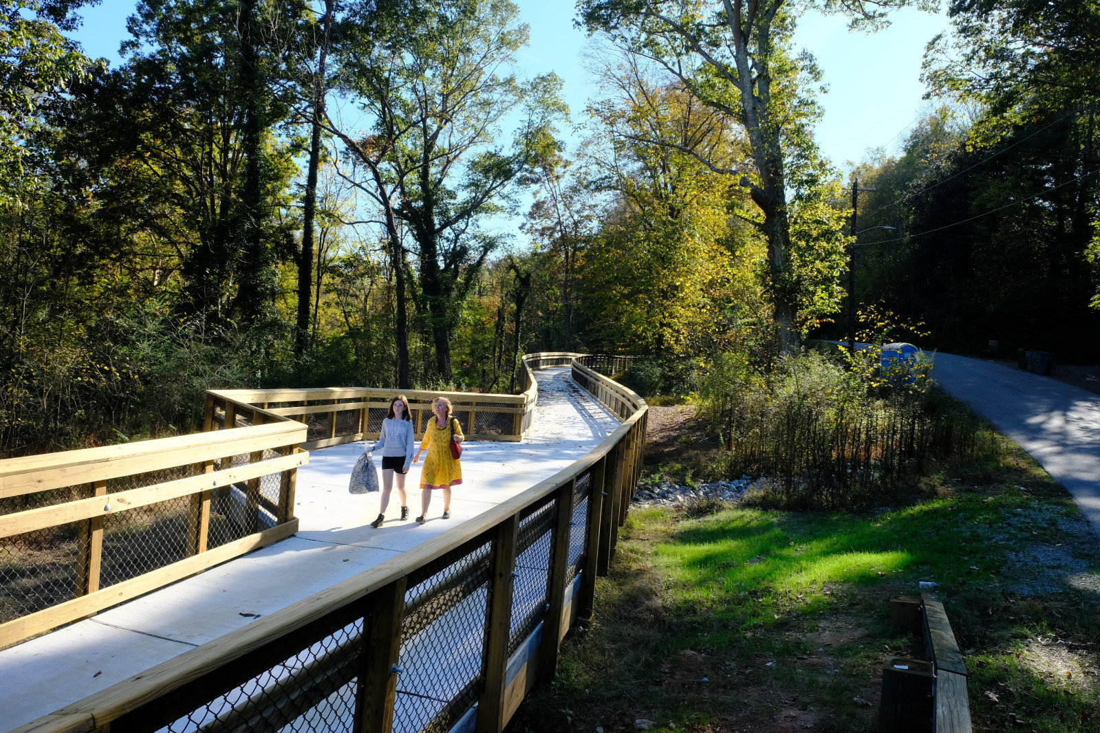 Two women walking on a wooden boardwalk in a wooded area, surrounded by trees and greenery.